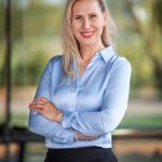 A smiling woman with long blonde hair, wearing a light blue blouse, stands with arms crossed against a blurred outdoor background.